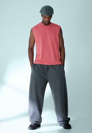 Maglietta senza maniche color corallo abbinata a pantaloni della tuta grigi larghi. L'outfit presenta una texture liscia, un design minimalista e scarpe in stile atletico.