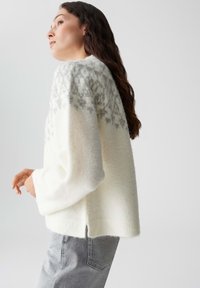 Maglione bianco in lana con una texture morbida e una rozza grigia decorata. Presenta maniche larghe e una vestibilità rilassata, abbinato a jeans grigi.