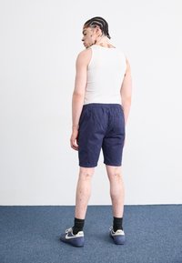 Débardeur blanc, shorts navy avec taille élastique et baskets navy avec accents crème. La peau avec des variations de pigmentation est visible.