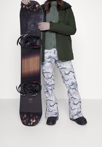 Snowboard con un design a gradiente nero e legno, dotato di attacchi neri. La persona indossa un abbigliamento esterno verde e pantaloni da neve grigi mimetici.