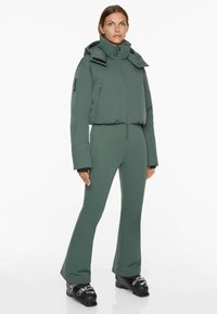 Green padded jacket with high collar and hood, paired with fitted green pants. Features zippers and textured fabric.