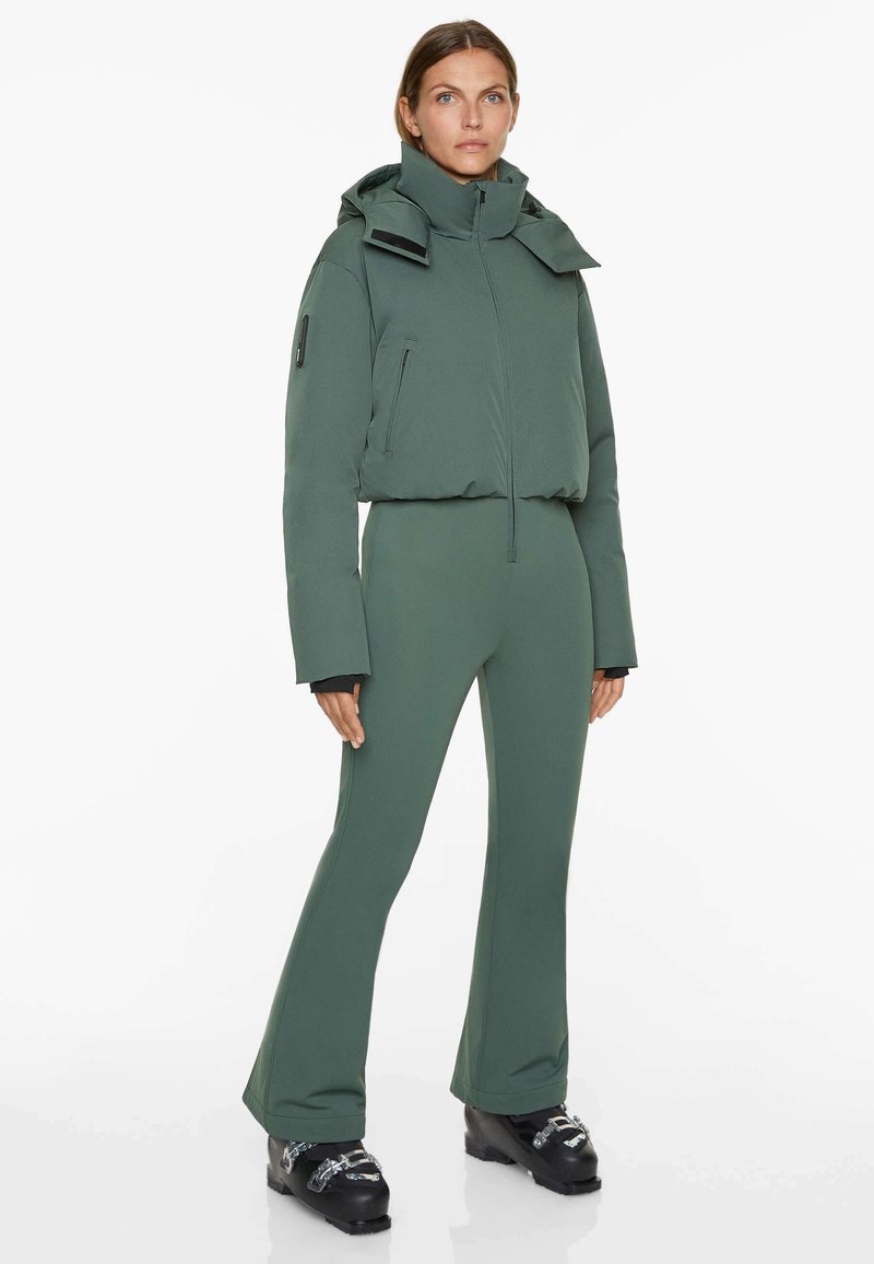 Green padded jacket with high collar and hood, paired with fitted green pants. Features zippers and textured fabric.