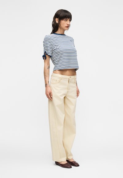 Ριγέ cropped τοπ με ναυτικές και λευκές ρίγες, δεμένα μανίκια· ανοιχτό μπεζ παντελόνια με φαρδύ πόδι και εμφανείς ραφές, συνδυασμένα με καφέ μπαλαρίνες.