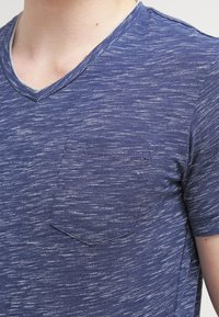 Detailní záběr na osobu oblečenou v modrém melírovaném triku s výstřihem do V a kapsou na levé straně hrudi.