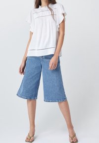 Blouse blanche avec des manches volantes et des accents brodés, associée à des culottes en denim bleu clair. Le mannequin porte des sandales vertes.
