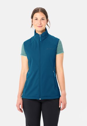 Frau trägt eine smaragdgrüne, ärmellose Weste mit Reißverschluss über einem kurzärmeligen grünen Shirt, steht mit entspannten Armen vor einem einfachen, hellen Hintergrund.