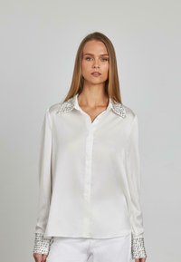 Blusa in satin bianco con colletto classico, arricchita da borchie argentate sul colletto e sui polsini, caratterizzata da una vestibilità morbida e maniche lunghe.