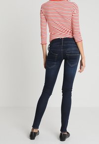 Woman wearing dark blue skinny jeans and a red-and-white striped long-sleeve shirt, standing with back to the viewer.