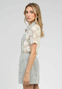 Blouse à manches courtes en dentelle florale, couleur crème, avec un col. Associée à une jupe grise texturée avec des boutons à l'avant et une coupe ajustée.