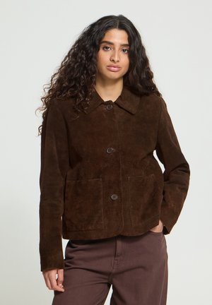Jeune femme aux longs cheveux bouclés portant une veste en suède marron foncé boutonnée et un pantalon assorti, debout, une main dans la poche.