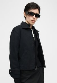 Άτομο με κοντά σκούρα μαλλιά που φοράει μαύρα γυαλιά ηλίου, μαύρο μπουφάν με φερμουάρ και μαύρο παντελόνι, σε απλό φωτεινό φόντο.