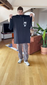 Juodas medvilnės T-marškinėlis su baltu grafinio spausdinimo ir prekės ženklo "Levi's" etikete priekyje. Modelis laiko marškinėlius šviesioje patalpoje.