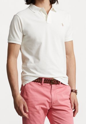 Mężczyzna w białej koszulce polo z krótkim rękawem, różowych spodniach, brązowym plecionym pasku i brązowym skórzanym zegarku na nadgarstku, stojący na jednolitym tle.