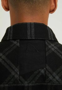 Černošedá kostkovaná košile s klopovým límcem, která má texturovanou tkaninu a malý trojúhelníkový štítek na zadní straně.