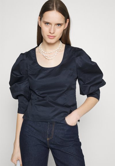 Ναυτικό μπλε, εφαρμοστό τοπ με φουσκωτά μανίκια, λεία υφή, χαμηλή στρογγυλή λαιμόκοψη, συνδυασμένο με σκούρα τζιν παντελόνια και στρωτή αλυσίδα με πέρλες.