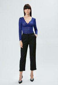 Blauwe kanten top met lange mouwen en scalloped halslijn, gecombineerd met zwarte op maat gemaakte broeken en zwarte hakken met decoratieve accenten.