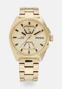 Reloj de oro con una pulsera metálica, caja octagonal, acentos en negro en la esfera y una esfera amarillo claro con subesferas y marcadores.