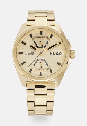 Reloj de oro con una pulsera metálica, caja octagonal, acentos en negro en la esfera y una esfera amarillo claro con subesferas y marcadores.