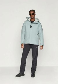 Veste isolée bleu clair avec capuche, accents noirs et fermeture éclair, associée à un pantalon noir et des chaussures noires. Modèle portant des lunettes de soleil.