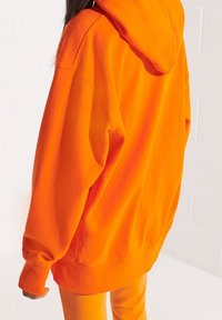 Sudadera de gran tamaño en color naranja con capucha ajustable y puños acanalados, hecha de una tela suave y texturizada. Combinada con pantalones naranja a juego.