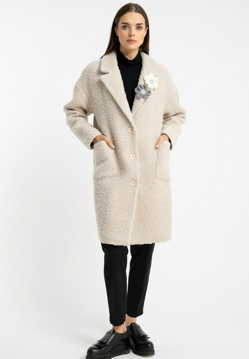 Jasno beżowy wełniany płaszcz o teksturowanym wykończeniu, z wykładanym kołnierzem, przednimi guzikami i dwoma bocznymi kieszeniami. Udekorowany kwiatowymi zdobieniami.