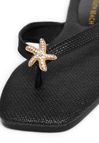 Infradito neri realizzati in materiale testurizzato, con un accento di stella marina dorata decorata con strass. Sottopiede di forma quadrata e design minimalista.