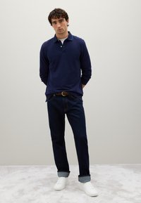 Mann mit lockigen Haaren, der ein navyblaues Langarm-Polo, dunkelblaue Jeans mit hochgerollten Säumen, einen braunen Gürtel und weiße Sneakers trägt, steht drinnen.