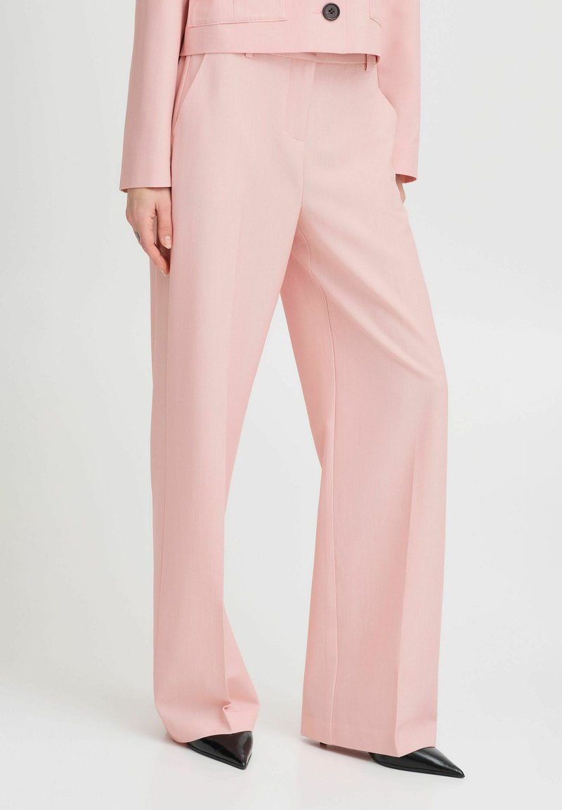 Lichtroze op maat gemaakte broek met een hoge taille en wijde pijpen. Heeft zijzakken en scherpe plooien, gecombineerd met puntige zwarte hakken.