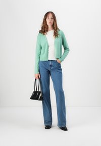 Cardigan verde-claro com decote redondo, usado sobre uma t-shirt branca, combinado com jeans boca-de-sino azuis. Uma bolsa preta e sapatos completam o visual.