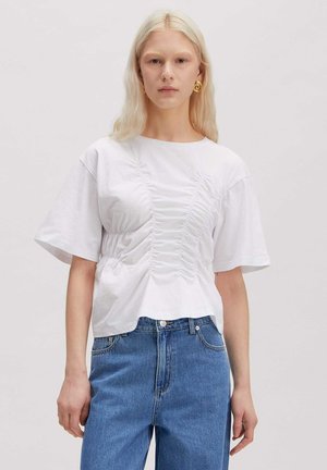 Blonde vrouw met gouden spiraalvormige oorbellen, een wit gerimpeld korte mouwen shirt en blauwe high-waist jeans tegen een effen achtergrond.