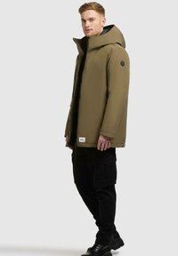 Olivgrüne gepolsterte Jacke mit großer Kapuze, schwarzem Reißverschluss, Seitentaschen und einem Logopatch nahe dem Saum, kombiniert mit schwarzen Cargo-Hosen und Stiefeln.