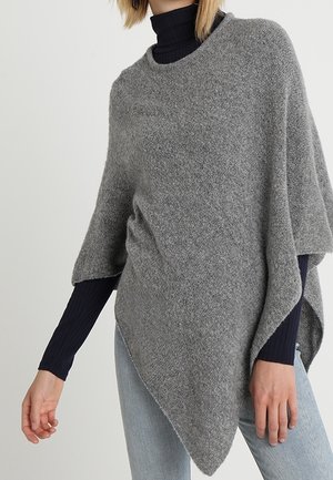 Kobieta ubrana w szary wełniany ponczo na czarnym golfie z długim rękawem i jasnoniebieskich jeansach, stoi z ramionami swobodnie opuszczonymi wzdłuż ciała.