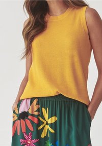 Kvinde iført en ærmeløs gul striktop og en grøn nederdel med farverig blomsterprint, hænderne i nederdelens lommer.