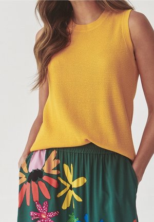 Kobieta w bezrękawnikowej, żółtej dzianinowej bluzce i zielonej spódnicy w kolorowy print kwiatowy, z rękami w kieszeniach spódnicy.