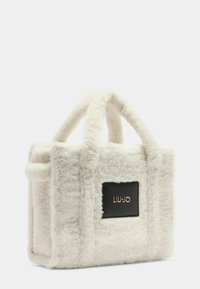 Borsa in peluche bianco testurizzato con manici corti e un badge con logo nero. Forma rettangolare con materiale morbido e dettagli di cucitura prominenti.