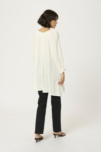 Witte lange mouw tuniek met een vloeiende ontwerp, gestructureerde stof en een ronde zoom, gecombineerd met zwarte strakke broek en zwarte sleehak sandalen.