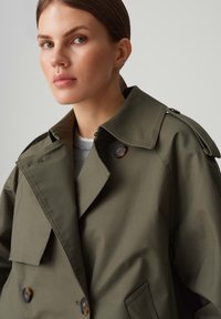 Kaki trenchcoat met een grote kraag, dubbele knoopsluiting en schouderstroken. Soepele stof met een lichte glans.