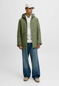 Grüne wasserdichte Jacke mit Kapuze, getragen über einem weiß gestreiften Poloshirt, kombiniert mit weit geschnittenen blauen Jeans und beigen Schuhen. Lässiger Streetwear-Stil.