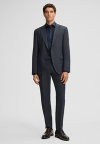 Donkergrijs op maat gemaakt pak met een gestructureerde afwerking, bijpassende broek en een donkerblauw overhemd. Voorzien van notch revers en knoopsluiting.