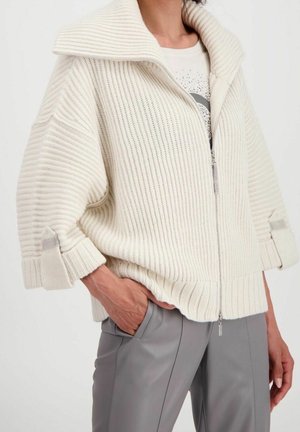 Chaqueta blanca de punto con cremallera y un amplio collar, de textura acanalada. Presenta puños doblados con un acento gris. Combinada con pantalones grises.
