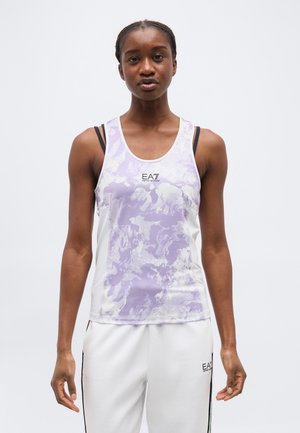 Žena oblečená v bezrukávovom športovom tielku s bielo-fialovým vzorom a bielych športových nohaviciach, stojaca pred jednofarebným bielym pozadím.