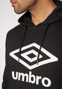 Čierna mikina s kapucňou s bielym diamantom a nápisom "umbro". Obsahuje kapucňu so sťahovacou šnúrkou a hladkú textúru látky.