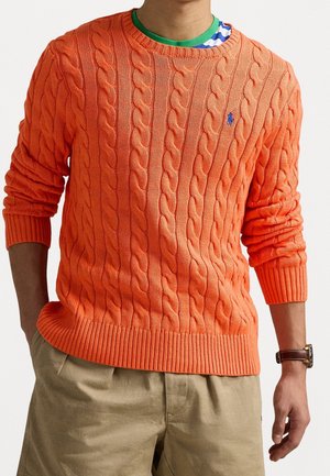 Person trägt einen orangefarbenen Zopfpullover über einem grün-weiß gestreiften Hemd, khakifarbene Hose und eine Armbanduhr mit braunem Lederarmband.