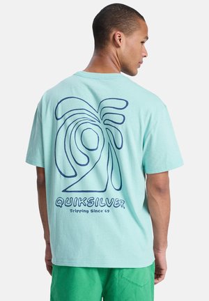 Mann trägt ein hellblaues T-Shirt mit abstraktem Wellenmuster und dem Schriftzug "QUIKSILVER Tripping Since '69" auf der Rückseite, kombiniert mit grünen Shorts.
