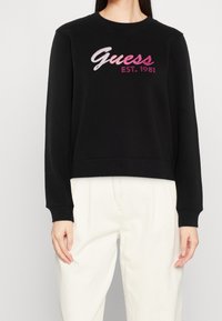 Felpa nera a maniche lunghe con la scritta "Guess EST. 1981" in testo rosa, indossata con pantaloni beige chiaro, tagliati al collo e a metà coscia.