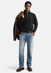 Zwarte sweater met rits, lichtblauwe jeans, bruine jas over de schouder, donkere schoenen met beige veters; casual outfit met minimale details.