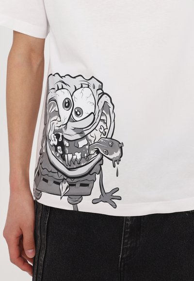 T-shirt blanc avec un motif d'un personnage de dessin animé aux traits du visage exagérés, des accents gris et une esthétique ludique et texturée.