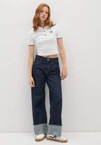 Polo shirt bianco a maniche corte con rifiniture e bottoni verdi, abbinato a jeans a vita alta blu scuro con pantaloni larghi e polsini in denim a contrasto.
