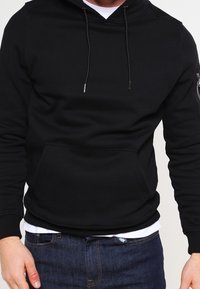 Černá mikina s kapucí s přední kapsou na brusle, stahovací kapucí a bočním zipem, vyrobená z hladké látky, kombinovaná s modrými džínami.