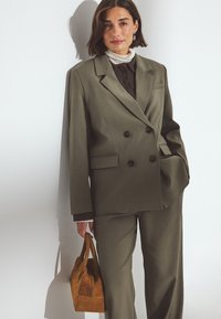 Zielona oliwkowa marynarka dwurzędowa z wyraźnymi ramionami i dużymi guzikami. W połączeniu z brązową koszulą zapinaną na guziki i trzymającą torebkę w kolorze jasnobrązowym.